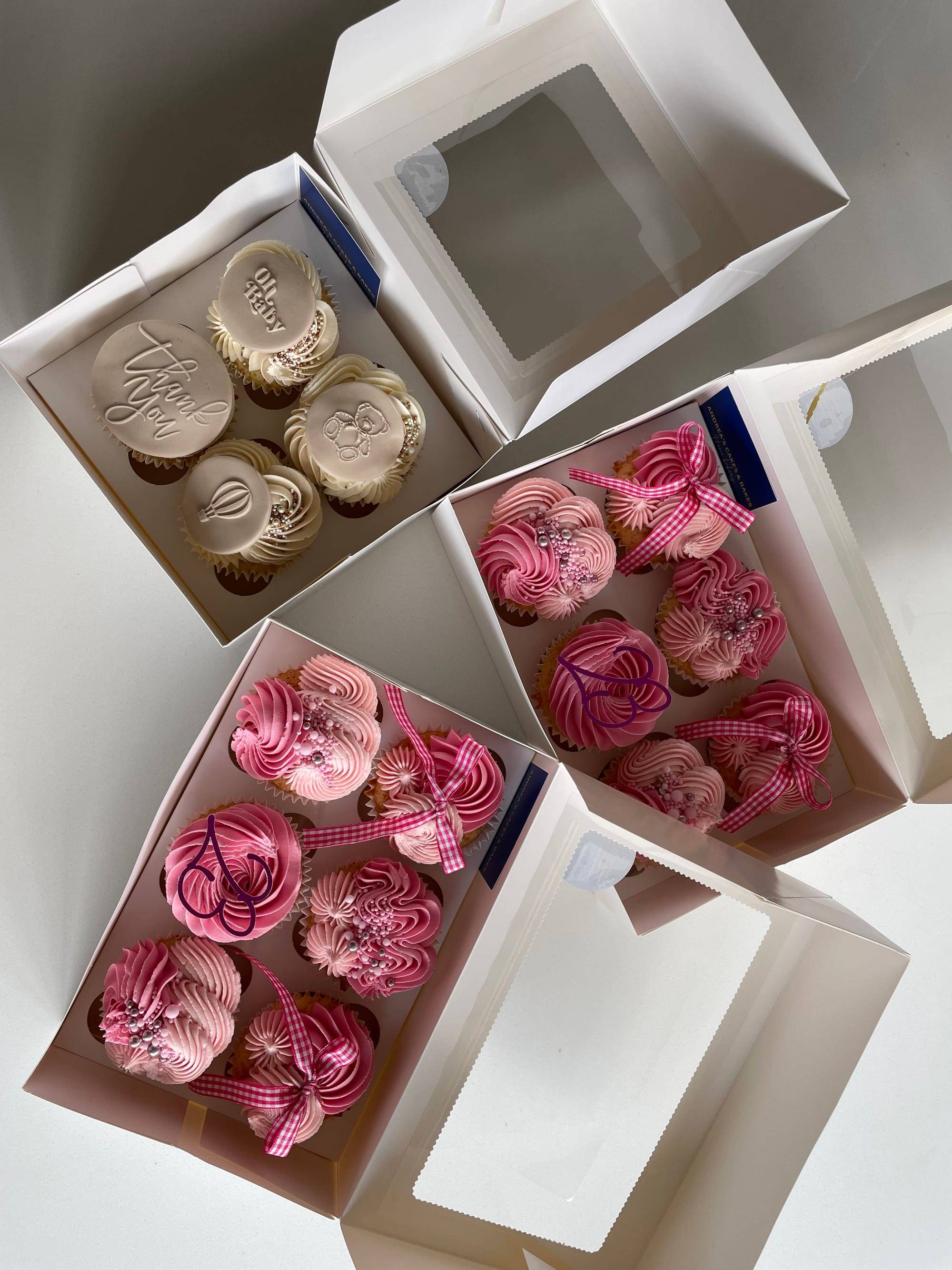 Assorted gift boxes of cupcakes, including ivory baby-shower designs and pink ribboned cupcakes