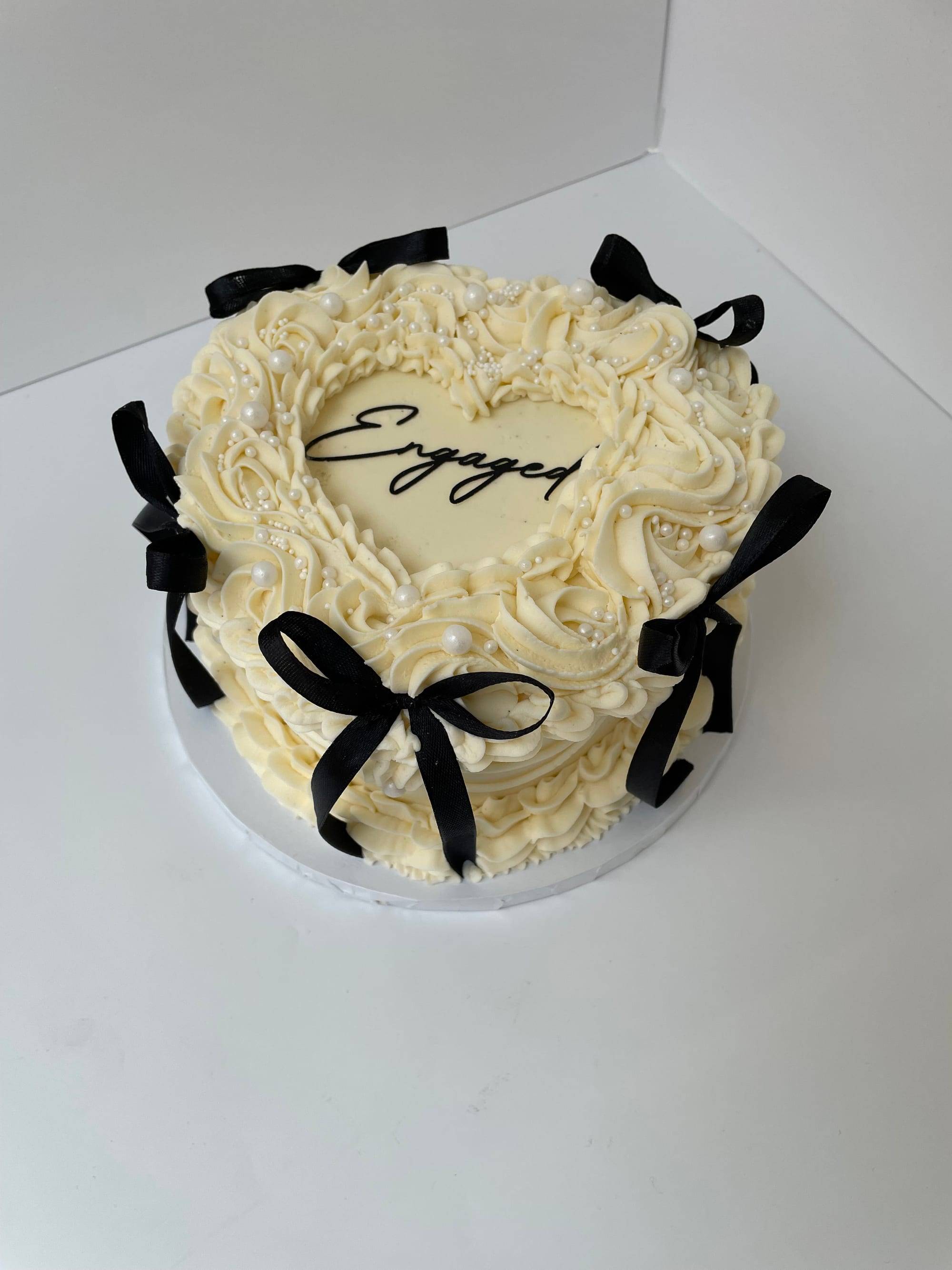 Heart-shaped engagement cake with ivory rosette buttercream, black ribbon bows and pearls, iced Engaged