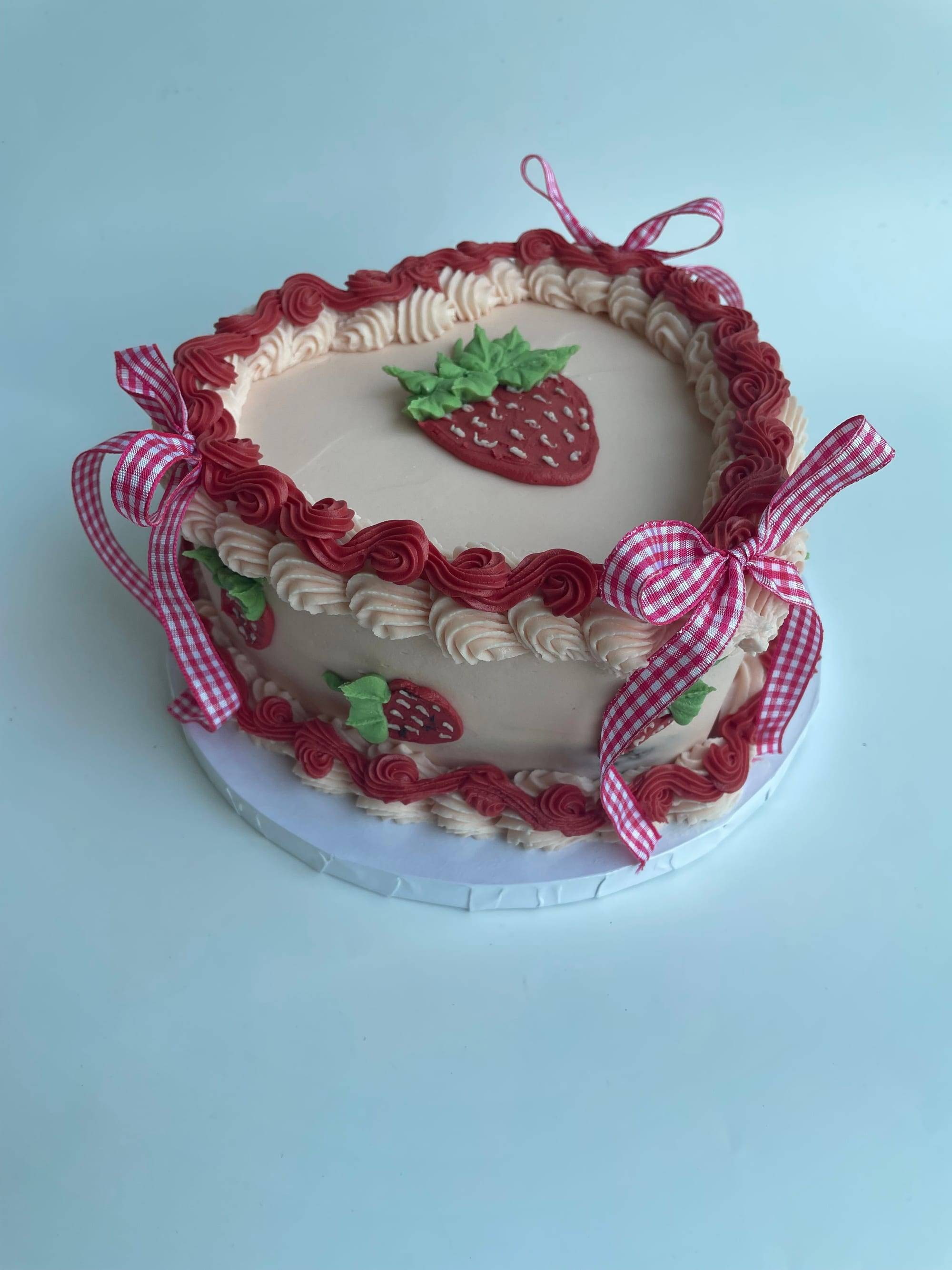 Heart-shaped cake decorated with piped strawberries on ivory buttercream and a red gingham ribbon